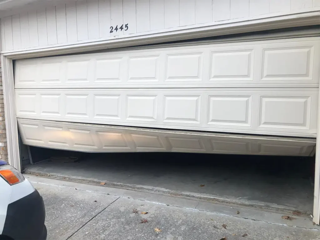 Garage door seriously damaged after falling off tracks, showing bent panels and displaced rollers on a residential garage door