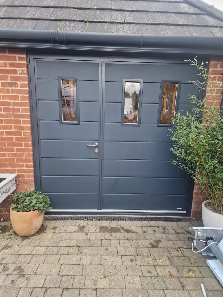 Grey side-hinged garage door with windows on a residential property, featuring a modern finish and glazed panels for natural light